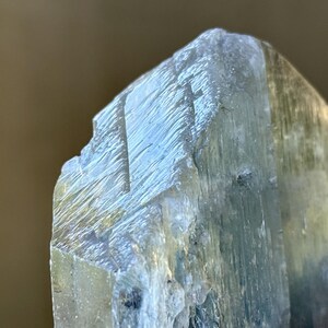 May include: Close-up of a rough-hewn, pale blue crystal formation. The crystal has a layered appearance with visible striations and a slightly translucent quality, suggesting a mineral specimen.