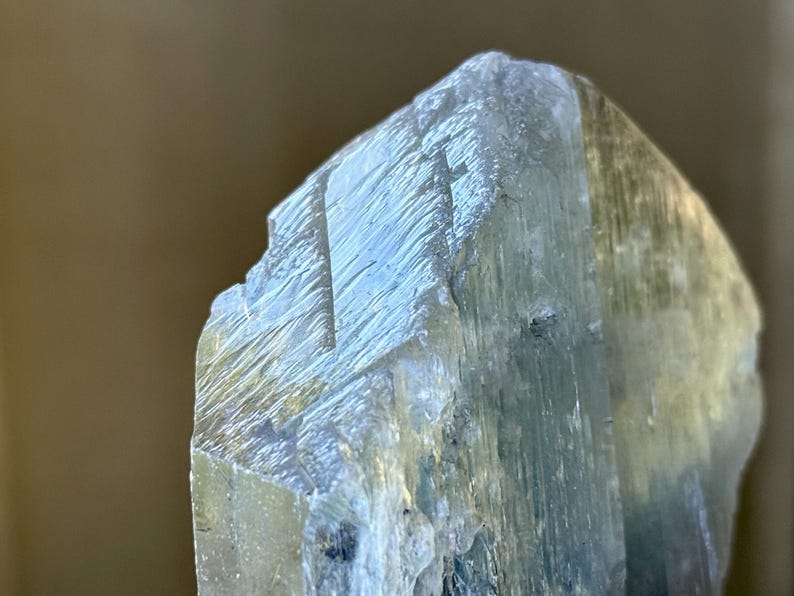 May include: Close-up of a rough-hewn, pale blue crystal formation. The crystal has a layered appearance with visible striations and a slightly translucent quality, suggesting a mineral specimen.