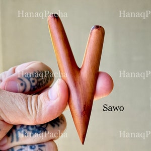 May include: A V-shaped wooden massage tool with a smooth, polished finish and natural reddish-brown wood grain. The object features two tapered prongs meeting at a single pointed base, with the text "HanaqPacha" and "Sawo" visible on the image.
