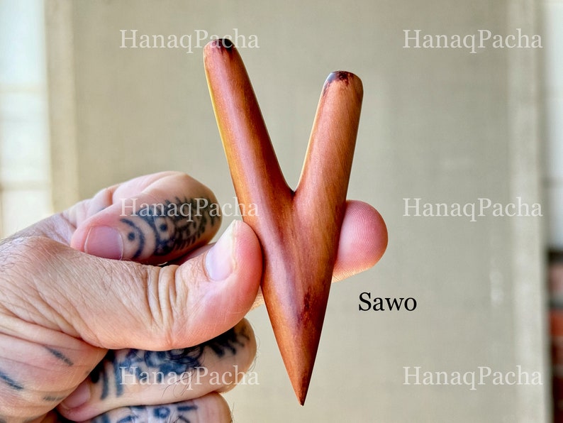 May include: A V-shaped wooden massage tool with a smooth, polished finish and natural reddish-brown wood grain. The object features two tapered prongs meeting at a single pointed base, with the text "HanaqPacha" and "Sawo" visible on the image.