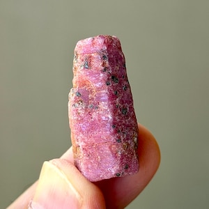 Könnte beinhalten: Ein roher, ungeschliffener Rubinkristall mit einem leuchtenden pink-roten Farbton. Der Edelstein hat eine hexagonale Säulenform mit einer rauen, strukturierten Oberfläche, natürlichen Facetten und kleinen dunklen Mineraleinschlüssen.