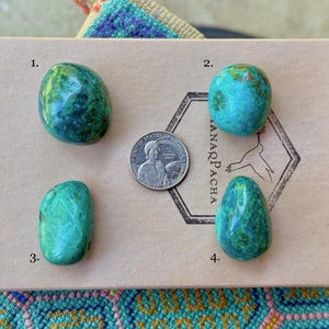 Puede incluir: Cuatro piedras pulidas en tonos verdes y azules se exhiben sobre una caja de cartón con una moneda para comparar el tamaño. Las piedras tienen una forma lisa y redondeada y patrones variados. La caja tiene el texto "ANARQPACHA" y un logotipo de pájaro.
