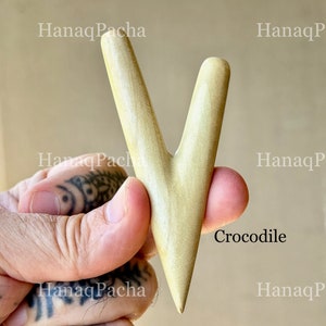 May include: A smooth, light-colored wooden massage tool with a V-shaped design featuring two tapered prongs of unequal length. The surface has a natural grain and a polished finish. The word Crocodile is printed in black text near the base.