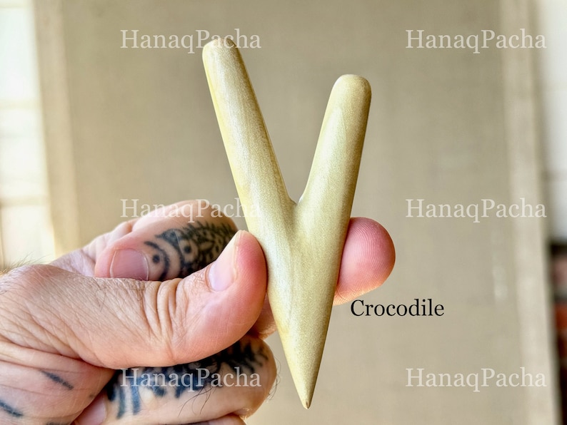 May include: A smooth, light-colored wooden massage tool with a V-shaped design featuring two tapered prongs of unequal length. The surface has a natural grain and a polished finish. The word Crocodile is printed in black text near the base.