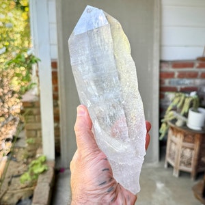 Può includere: Un grande cristallo di quarzo trasparente con una punta appuntita e una superficie ruvida e naturale. Il cristallo è tenuto in una mano, mostrando le sue dimensioni e la sua chiarezza.