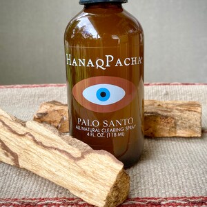 Peut inclure: Une bouteille en verre brun avec une &eacute;tiquette blanche qui indique "HANAPACHA" et "PALO SANTO ALL NATURAL CLEARING SPRAY 4 FL. OZ. (118 ML)". L'&eacute;tiquette pr&eacute;sente &eacute;galement un symbole d'&oelig;il bleu sur fond brun. La bouteille est pos&eacute;e sur un morceau de bois.