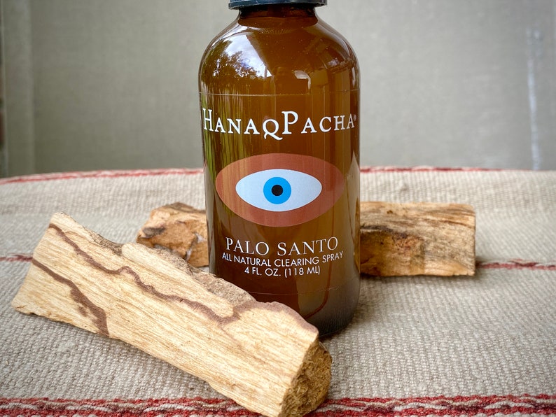 Peut inclure: Une bouteille en verre brun avec une &eacute;tiquette blanche qui indique "HANAPACHA" et "PALO SANTO ALL NATURAL CLEARING SPRAY 4 FL. OZ. (118 ML)". L'&eacute;tiquette pr&eacute;sente &eacute;galement un symbole d'&oelig;il bleu sur fond brun. La bouteille est pos&eacute;e sur un morceau de bois.