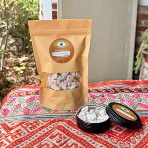 May include: A brown paper bag containing white copal resin is displayed on a patterned textile. A small, open black tin with resin and a closed tin with a label are also present. The label reads "HanaqPacha" and "Oaxacan White Copal".