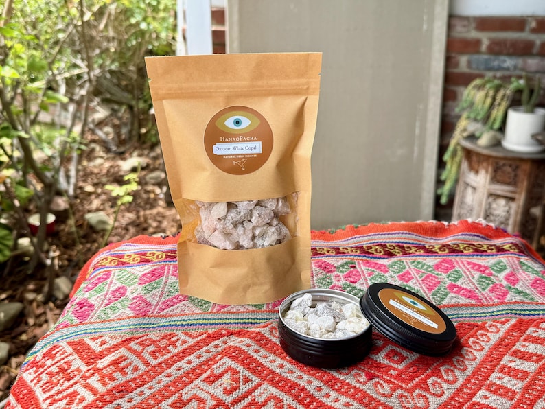 May include: A brown paper bag containing white copal resin is displayed on a patterned textile. A small, open black tin with resin and a closed tin with a label are also present. The label reads "HanaqPacha" and "Oaxacan White Copal".