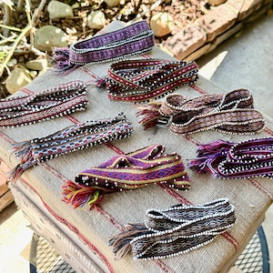 May include: A collection of colorful woven belts with intricate patterns and fringed ends. The belts feature various colors, including purple, red, and brown, and are displayed on a patterned fabric surface.