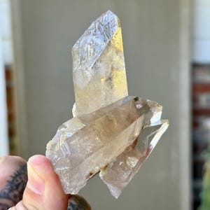 May include: A cluster of clear quartz crystals with a light brown hue. The crystals have sharp, pointed edges and a textured surface. The quartz is held in a hand, with a blurred background of a building and greenery.