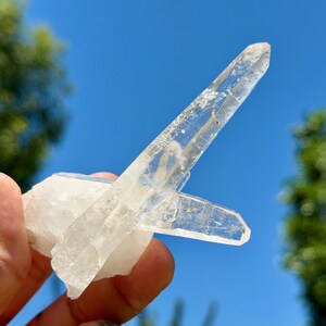 May include: A clear quartz crystal cluster with multiple points, held against a bright blue sky. The crystal is transparent with visible internal structures and imperfections. The natural light highlights the crystal's facets and textures.