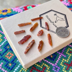 Puede incluir: Una colección de cristales en bruto de color ámbar dispersos sobre una caja beige, junto a una moneda. La caja tiene un logotipo con el texto "HanaqPacha" y una ilustración de un pájaro. Los cristales varían en tamaño y forma.
