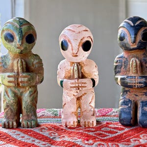 May include: Three carved alien figurines in green, pink, and blue-black hues. Each figure has large oval eyes and clasped hands. The figures are displayed on a patterned red and white textile.