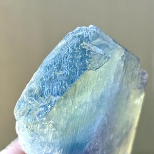 May include: Close-up of a raw, natural fluorite crystal. The crystal displays a gradient of colors, including shades of blue, green, and a hint of purple. The surface has a rough, textured appearance, with visible crystalline structures.