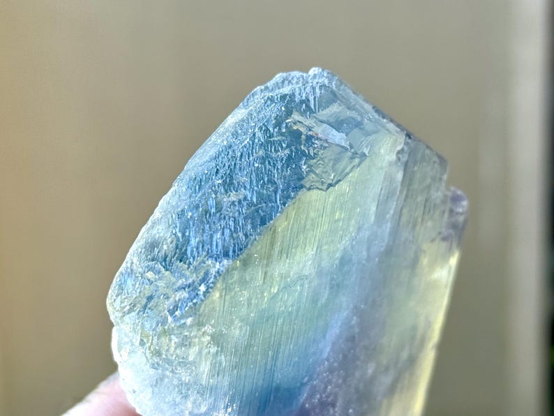 May include: Close-up of a raw, natural fluorite crystal. The crystal displays a gradient of colors, including shades of blue, green, and a hint of purple. The surface has a rough, textured appearance, with visible crystalline structures.