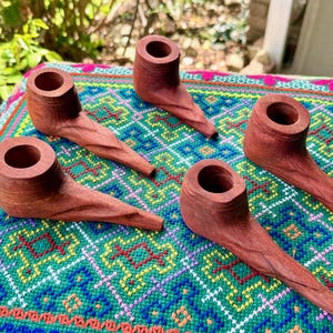 May include: Five hand-carved wooden pipes with a reddish-brown finish. The pipes are displayed on a colorful, patterned textile. Each pipe has a bowl and a long, slender stem, showcasing the natural wood grain.