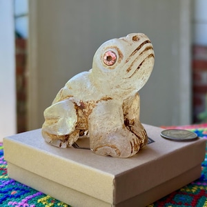 Puede incluir: Una figura de rana translúcida con un ojo rosado se encuentra sobre una caja beige. La rana tiene detalles y una superficie texturizada. Una moneda descansa sobre la caja junto a la rana. El fondo está borroso.