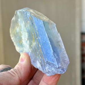 May include: A translucent, blue-hued crystal with a layered appearance. The crystal has a rough, natural texture and is held in a hand. The crystal's color shifts from light blue to a deeper shade, with hints of yellow.