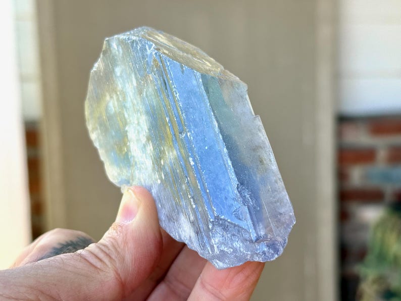 May include: A translucent, blue-hued crystal with a layered appearance. The crystal has a rough, natural texture and is held in a hand. The crystal's color shifts from light blue to a deeper shade, with hints of yellow.