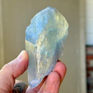 May include: A large, raw aquamarine crystal with a pale blue hue. The gemstone is held in a hand, showcasing its natural, irregular shape and translucent quality. The crystal's surface reflects light, highlighting its crystalline structure.