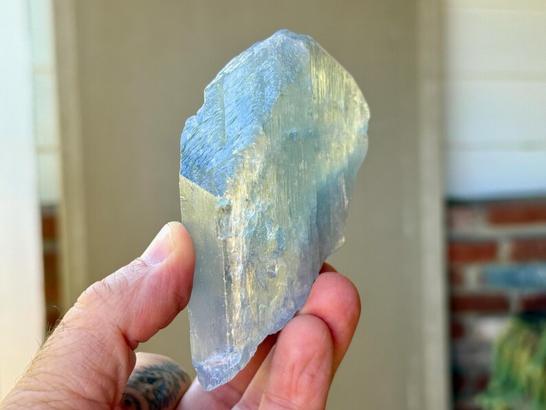 May include: A large, raw aquamarine crystal with a pale blue hue. The gemstone is held in a hand, showcasing its natural, irregular shape and translucent quality. The crystal's surface reflects light, highlighting its crystalline structure.