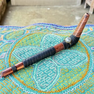 May include: A handcrafted wooden pipe with a curved design. The pipe features a dark brown wooden stem wrapped in black leather and a lighter wood bowl. A decorative stone is set into the leather. The pipe rests on a colorful patterned textile.