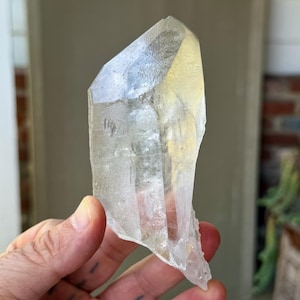 Puede incluir: Un cristal de cuarzo transparente con una punta y una superficie rugosa y texturizada. El cristal se sostiene en una mano, mostrando su apariencia natural y translúcida. El cristal es de color claro y neutro.