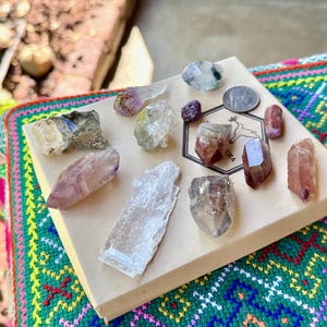 Puede incluir: Una colección de varios cristales y piedras preciosas en tonos morados, transparentes y marrones, dispuestos sobre una caja beige. La caja está colocada sobre un textil colorido y estampado. La imagen incluye una moneda y un pequeño logotipo con las letras "CHA".