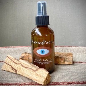 Peut inclure: Une bouteille en verre brun avec un vaporisateur noir et une &eacute;tiquette blanche qui dit "HANAQ PACHA" et "PALO SANTO ALL NATURAL CLEARING SPRAY 4 FL. OZ. (118 ML)". L'&eacute;tiquette pr&eacute;sente &eacute;galement un &oelig;il bleu avec une pupille blanche sur fond rouge. La bouteille est pos&eacute;e sur une surface ray&eacute;e brun et blanc avec deux morceaux de bois brun.