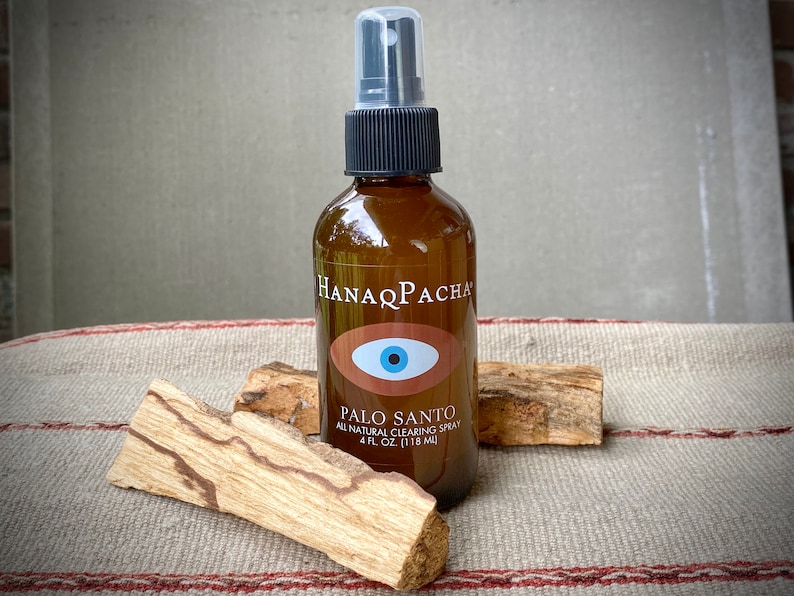 Peut inclure: Une bouteille en verre brun avec un vaporisateur noir et une &eacute;tiquette blanche qui dit "HANAQ PACHA" et "PALO SANTO ALL NATURAL CLEARING SPRAY 4 FL. OZ. (118 ML)". L'&eacute;tiquette pr&eacute;sente &eacute;galement un &oelig;il bleu avec une pupille blanche sur fond rouge. La bouteille est pos&eacute;e sur une surface ray&eacute;e brun et blanc avec deux morceaux de bois brun.