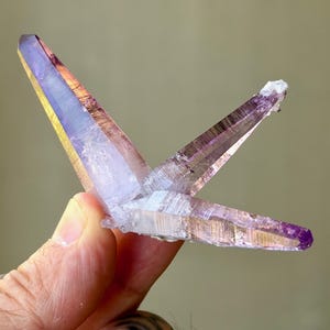 May include: A cluster of three translucent amethyst crystal points with a distinct purple hue. The crystals are elongated and pointed, featuring natural facets and internal inclusions that catch the light, creating a gradient of light and dark violet tones.
