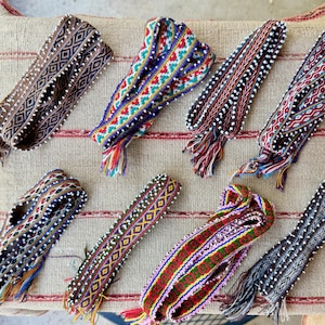 May include: A collection of eight woven belts with intricate patterns and colorful designs. The belts feature various color combinations, including brown, blue, red, and green, with decorative tassels at the ends. They are displayed on a textured, neutral-toned surface.