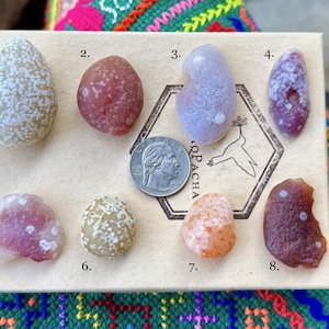 May include: An assortment of eight polished stones in various shapes and colors, including white, red, purple, and orange. A quarter coin is placed next to the stones for size comparison. The stones are arranged on a light brown surface.