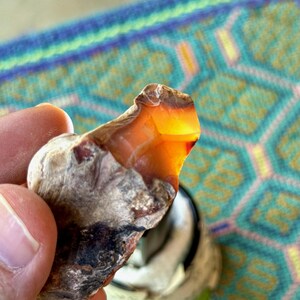 May include: A close-up of a rough, brown, and gray agate stone with a bright orange center. The stone is being held in a hand.
