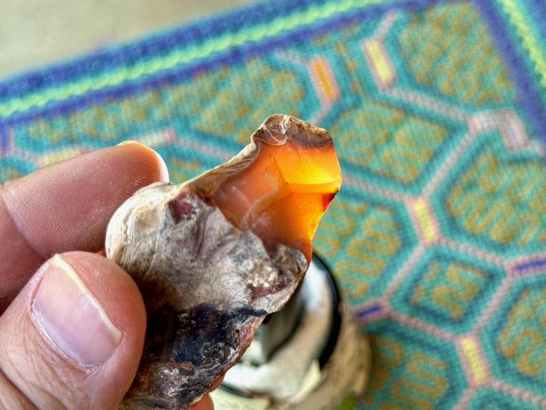 May include: A close-up of a rough, brown, and gray agate stone with a bright orange center. The stone is being held in a hand.