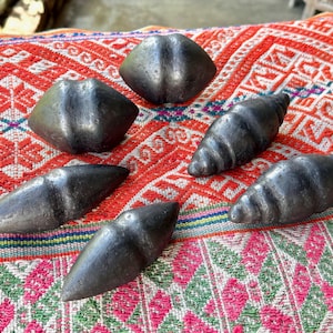 Puede incluir: Un surtido de objetos de color gris oscuro, parecidos a piedras. La colección incluye formas geométricas y piezas en espiral, dispuestas sobre un textil vibrante con patrones en tonos rojos, blancos y verdes. Los objetos parecen estar tallados a mano.