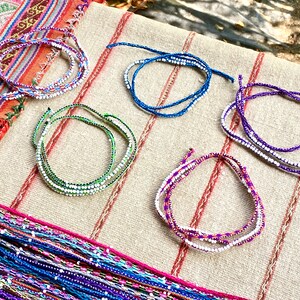 K&ouml;nnte beinhalten: F&uuml;nf bunte Perlenarmb&auml;nder mit wei&szlig;en Perlen auf einem beigefarbenen Stoffhintergrund. Die Armb&auml;nder bestehen aus F&auml;den in verschiedenen Farben, darunter Lila, Blau, Gr&uuml;n und Pink.