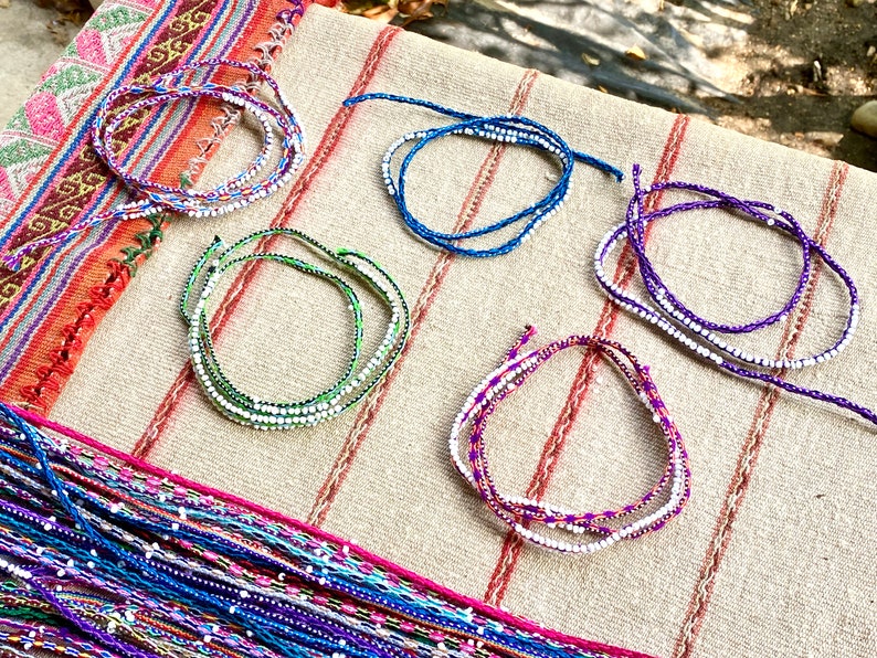 K&ouml;nnte beinhalten: F&uuml;nf bunte Perlenarmb&auml;nder mit wei&szlig;en Perlen auf einem beigefarbenen Stoffhintergrund. Die Armb&auml;nder bestehen aus F&auml;den in verschiedenen Farben, darunter Lila, Blau, Gr&uuml;n und Pink.