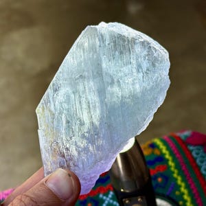 May include: A translucent, pale blue crystal with a rough, uneven surface. The crystal is held in a hand, with a blurred background. The crystal's texture appears to have vertical striations. The overall theme is related to minerals and crystals.