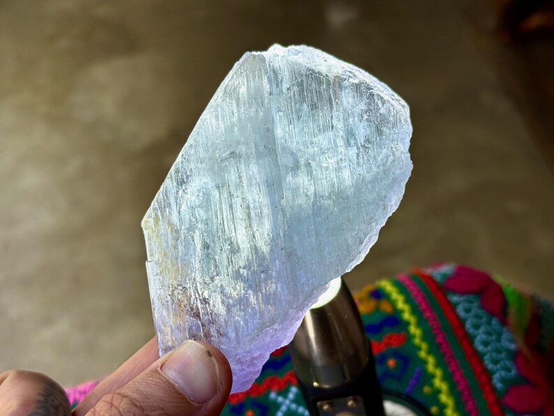 May include: A translucent, pale blue crystal with a rough, uneven surface. The crystal is held in a hand, with a blurred background. The crystal's texture appears to have vertical striations. The overall theme is related to minerals and crystals.