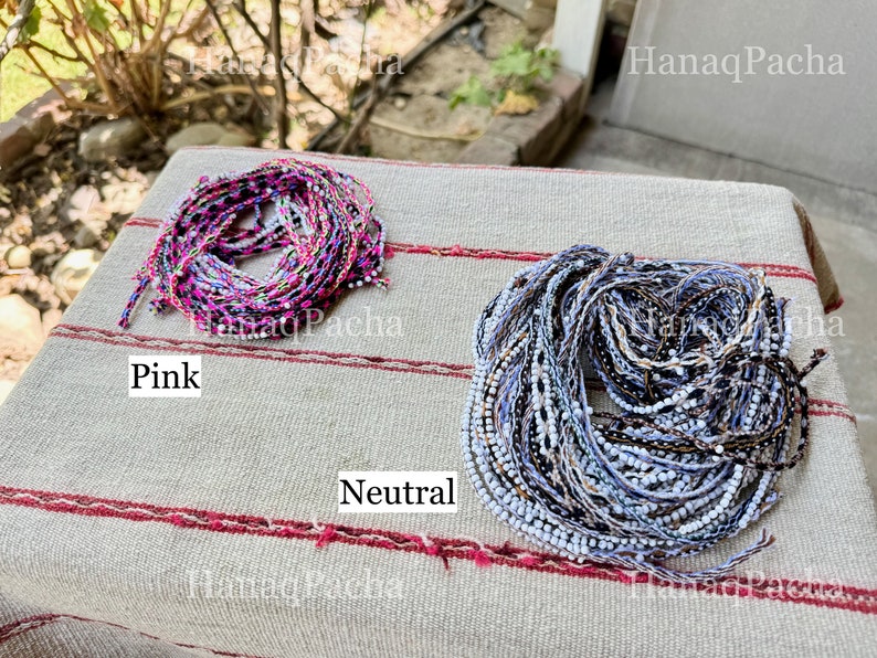 K&ouml;nnte beinhalten: Zwei Spulen mit farbigem Garn, eine rosa und eine neutral, auf einer gestreiften Stoffoberfl&auml;che. Der Text "Hanaq Pacha" ist im Bild sichtbar.