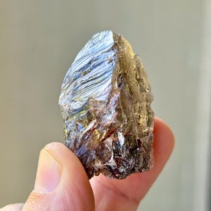May include: A raw, metallic-looking mineral specimen with a crystalline structure. The stone has a dark, reflective surface with shades of silver and brown. The mineral is held in a hand, showcasing its natural, unpolished form.