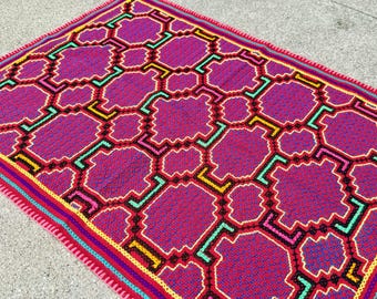 May include: A vibrant, handmade crochet blanket featuring a geometric pattern in shades of fuchsia, red, black, yellow, and teal. The blanket has a detailed border with multiple color stripes.