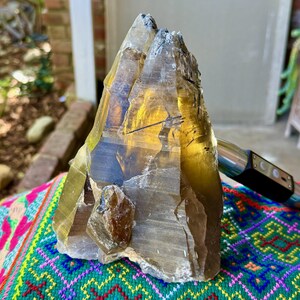 以下が含まれることがあります： 金茶色の大きな原石のスモーキークォーツの結晶。 結晶はざらざらした質感の表面を持ち、カラフルな模様の織物の上に展示されています。 銀と黒のデバイスが部分的に見えています。