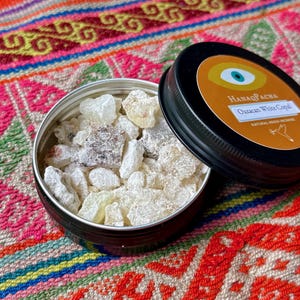 May include: A small, open black tin filled with white copal resin. The lid, also black, is partially open, revealing the product name "Oaxacan White Copal." The background is a colourful textile with a geometric pattern.