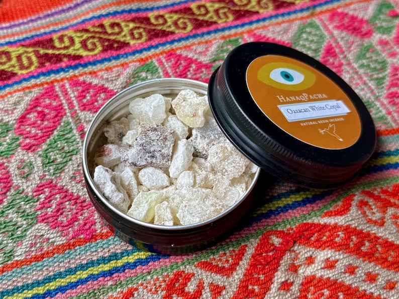 May include: A small, open black tin filled with white copal resin. The lid, also black, is partially open, revealing the product name "Oaxacan White Copal." The background is a colourful textile with a geometric pattern.