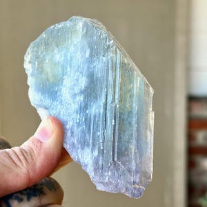 May include: A translucent, light blue crystal with a rough, textured surface. The crystal has vertical striations and a subtle gradient of color, transitioning from a pale blue to a slightly darker hue. The crystal is held in a hand.