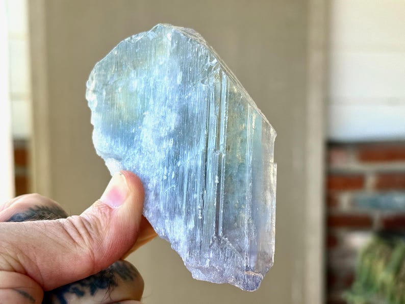 May include: A translucent, light blue crystal with a rough, textured surface. The crystal has vertical striations and a subtle gradient of color, transitioning from a pale blue to a slightly darker hue. The crystal is held in a hand.