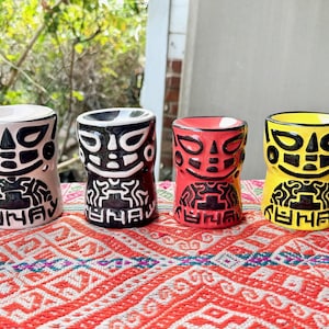Puede incluir: Cuatro tazas tiki de cerámica coloridas. Cada taza presenta un diseño de rostro estilizado con detalles en negro. Las tazas son de color rosa, negro, rojo y amarillo, y están dispuestas sobre un textil estampado.